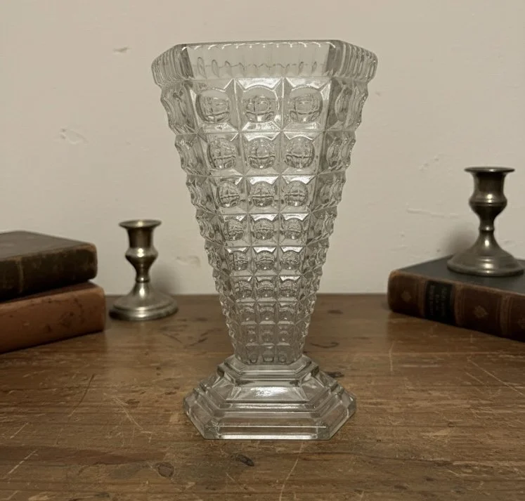 A vintage cut glass vase with a geometric pattern on a wooden surface, flanked by two small silver candlestick holders with books in the background.