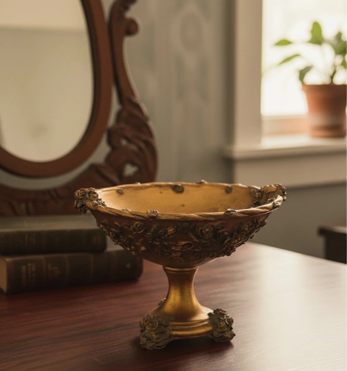 An ornate gold-colored decorative bowl with detailed floral designs, sitting on a wooden surface with a mirror and books in the background.