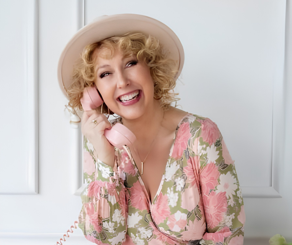 A woman with curly blonde hair wearing a light-colored hat and floral dress, smiling, holding a pink vintage phone receiver.