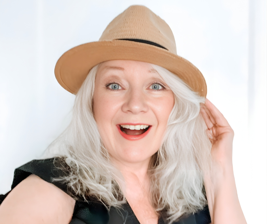 A woman with long gray hair wearing a tan hat and a black sleeveless top, smiling and touching her hat.