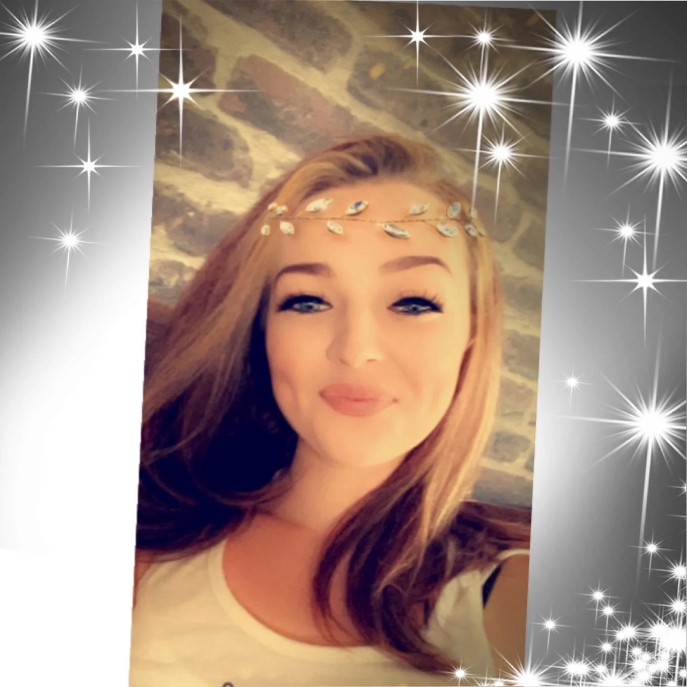 A young woman with light skin and long red hair smiling at the camera, wearing a decorative headband with leaf-shaped embellishments, with a background of a brick wall and celestial sparkles overlay.