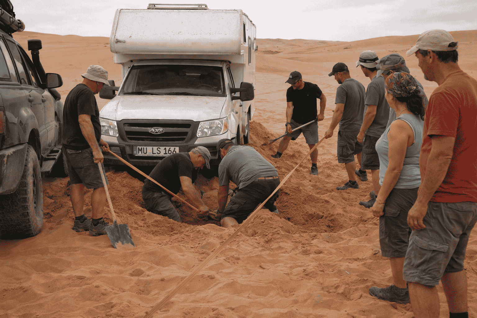 Caravan Weltreise Gruppe versuchen, ein Auto in der Wüste aus dem Sand zu ziehen, mit Schaufeln und Seilen.