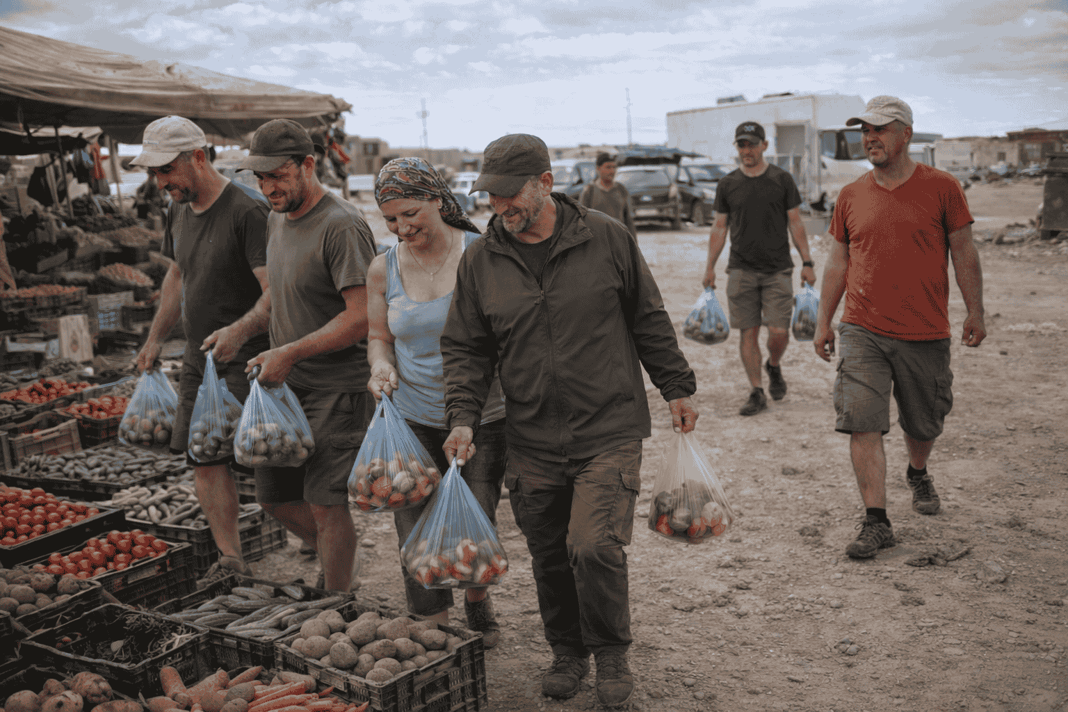 Caravan Weltreise Reisegruppe  beim Einkauf auf einem Outdoor-Markt, die verschiedene Gemüsesorten in Plastiktüten tragen, mit Ständen im Hintergrund.