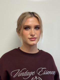 Inhaberin von Caravan Weltreisen Junge Frau mit blonden Haaren, trägt eine burgunderfarbene Bluse mit weißen Schriftzug 'Vintage Essence'.