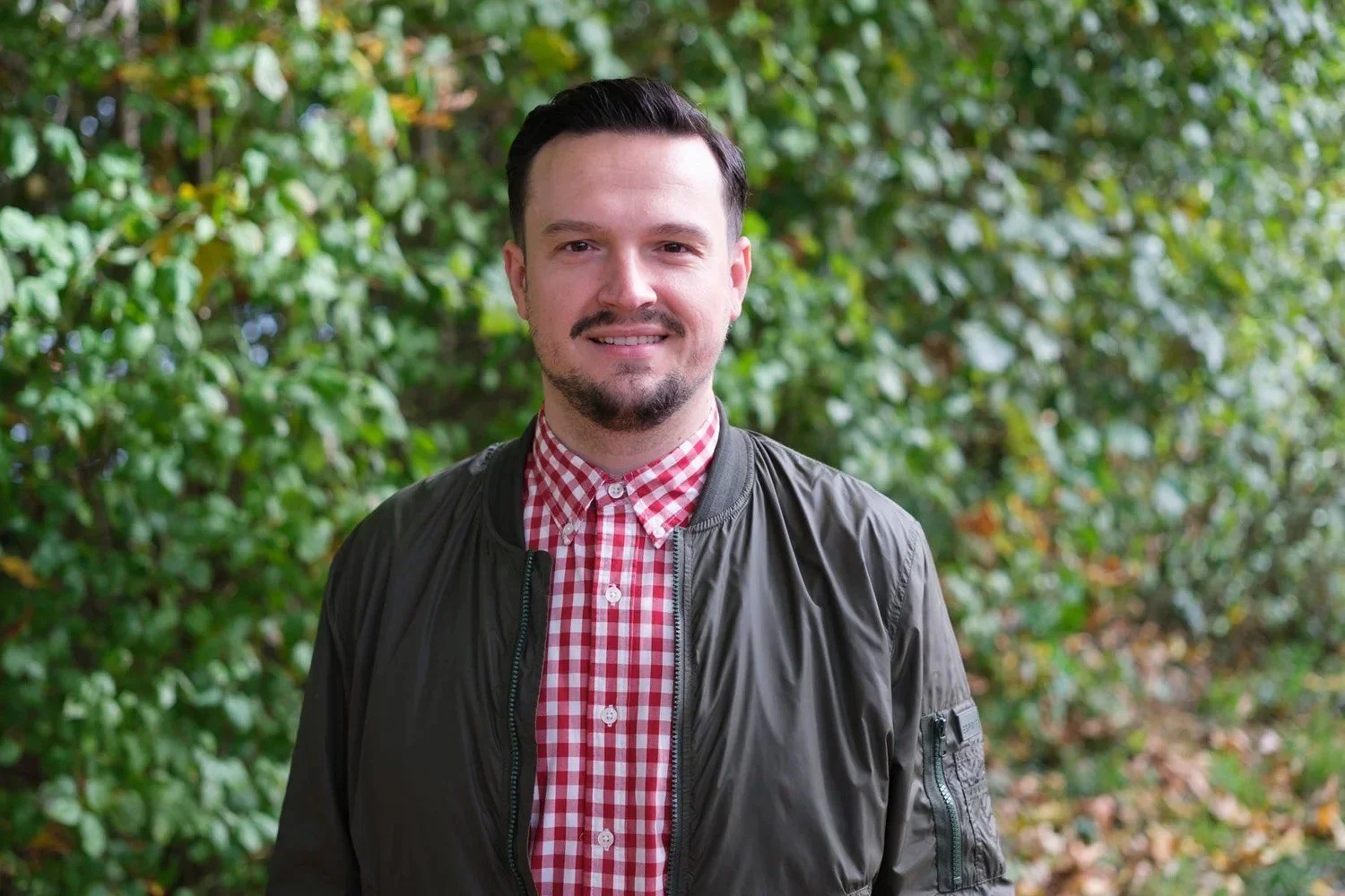 Junger Mann im Freien vor grünen Büschen, trägt eine schwarze Jacke und ein rotes kariertes Hemd.