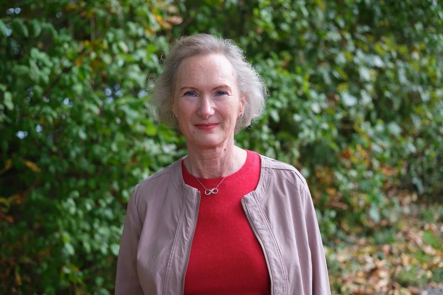 Eine ältere Frau mit grauem Haar steht vor einem grünen Busch, trägt eine beige Jacke, ein rotes Oberteil und ein Infinity-Schmuck. Sie lächelt leicht und schaut in die Kamera.