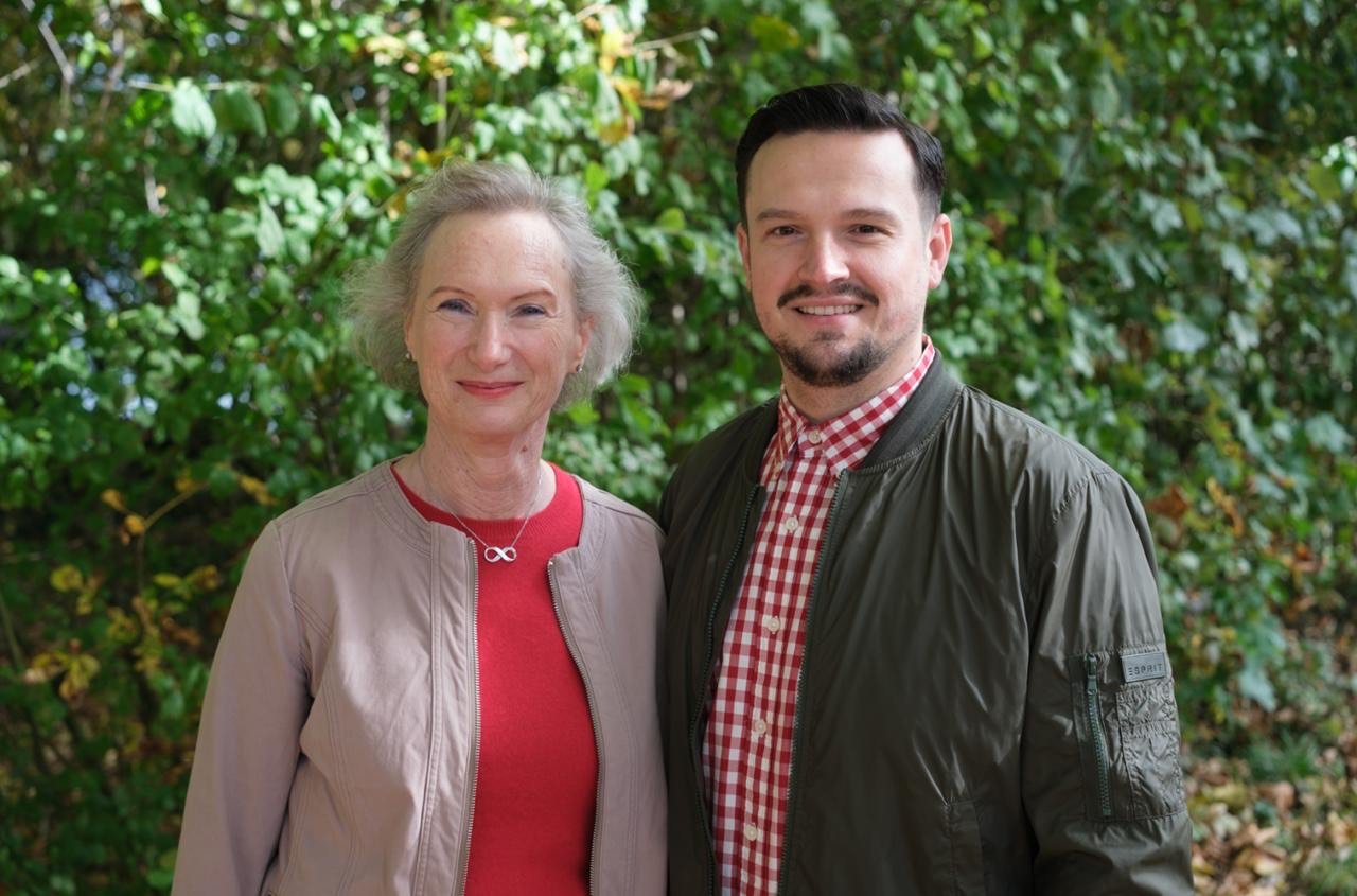 Ein älterer und ein jüngerer Mann stehen nebeneinander vor einer grünen Laube. Beide lächeln in die Kamera, sie trägt einen beige Jacke und ein rotes Oberteil, er trägt ein schwarzes Jacke und ein rotes Karohemd.