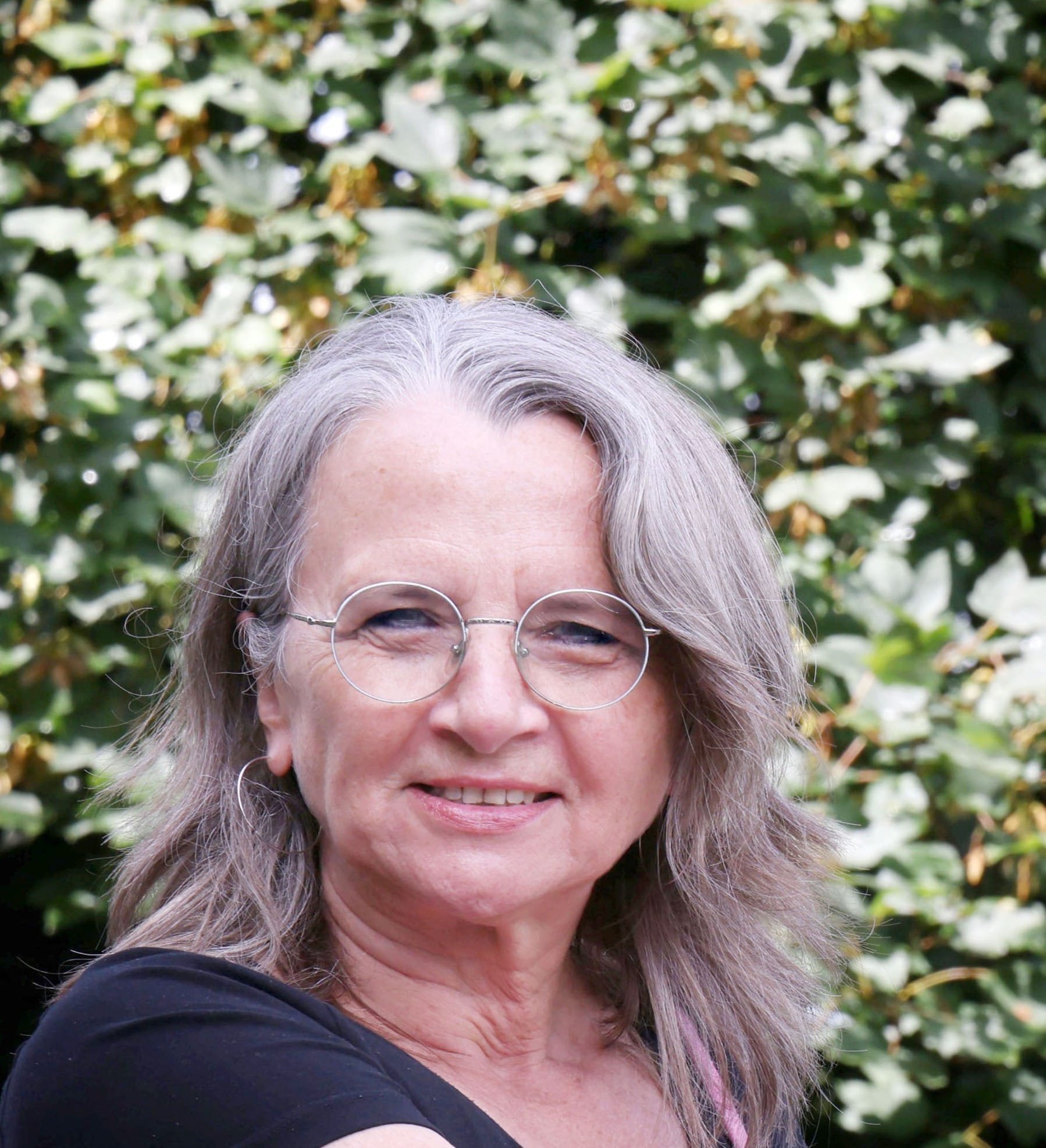 Porträt einer älteren Frau mit grauen Haaren und Brille, lächelnd vor einer grünen Laubbäume im Hintergrund.