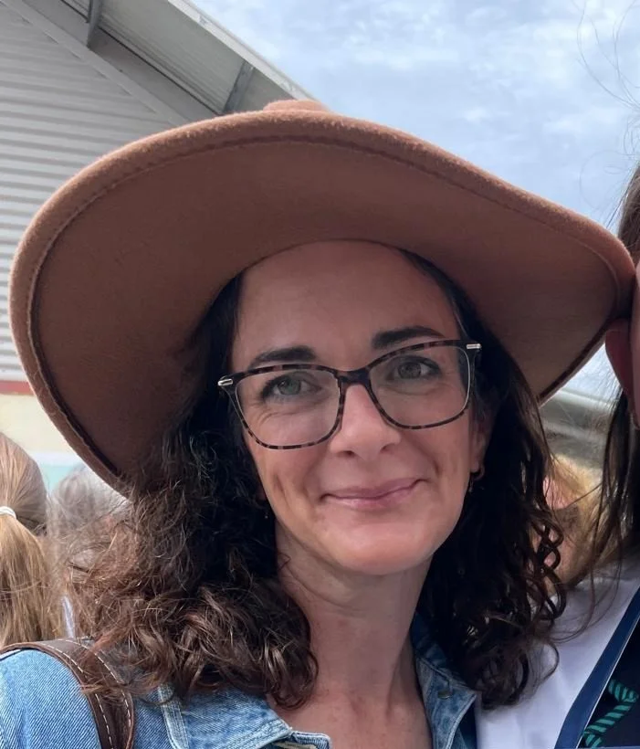 A woman with glasses, curly brown hair, and a wide-brimmed hat smiling outdoors.