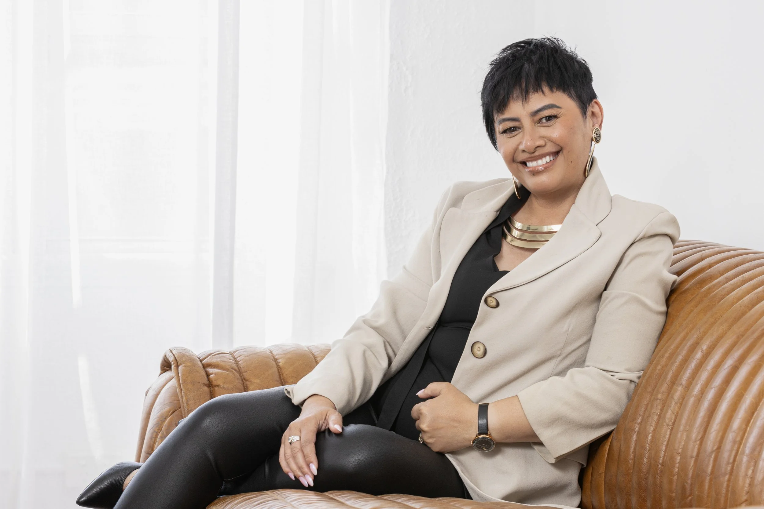 A woman with short black hair, wearing a beige blazer, black top, and leather pants, sitting on a brown leather couch with a white background, smiling at the camera.