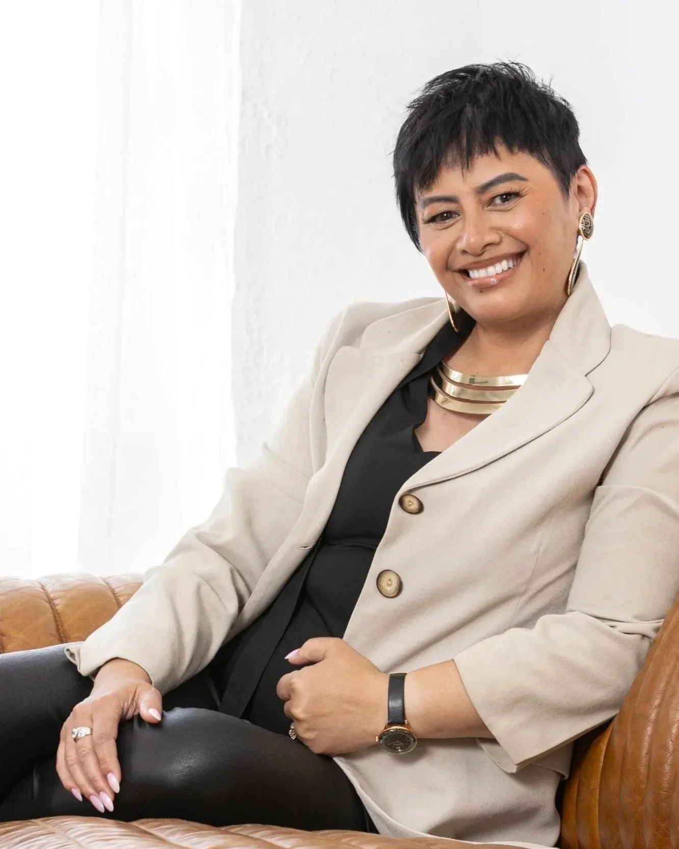 A woman with short dark hair, wearing a beige blazer and black top, sitting on a brown leather couch, smiling, with jewelry including earrings, a necklace, a watch, and a ring.