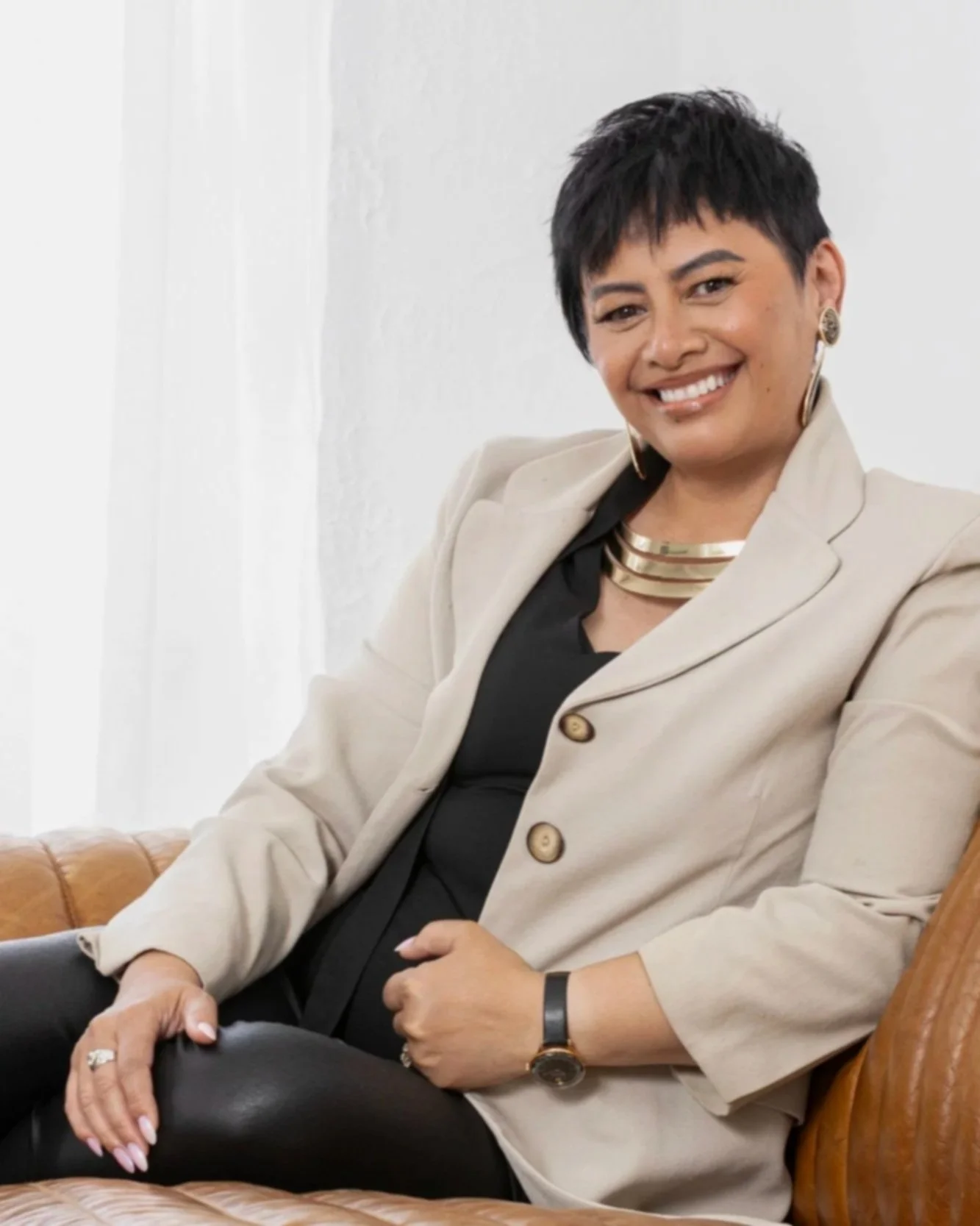 A smiling woman with short black hair, wearing a beige blazer, black top, gold jewelry, and a watch, sitting on a brown leather chair in a bright room.