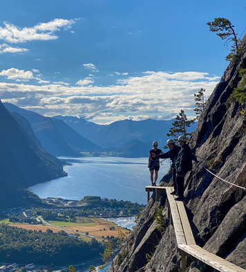 Tre personer går via ferrata i Åndalsnes, med fjell og fjord i bakgrunnen under en klar blå himmel.