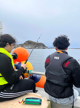 To personer fisker fra en båt med bøyer i vannet, mot en øy i Bakgrunnen.