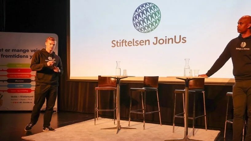 To personer er på en scene med to små bord og tre stoler. Bak en stor skjerm med teksten "Stiftelsen JoinUs" og logo. Den ene personen står ved det høyre bord, den andre er til venstre, begge er kledd i mørke klær.