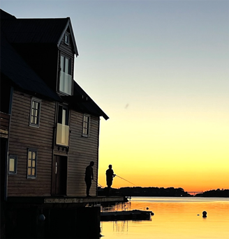 To personer står på en brygge ved vannet mot en solnedgang, med en stor svart trebåt i bakgrunnen. Bilde fra Drømmebrygga i regi av Stiftelsen JoinUs.