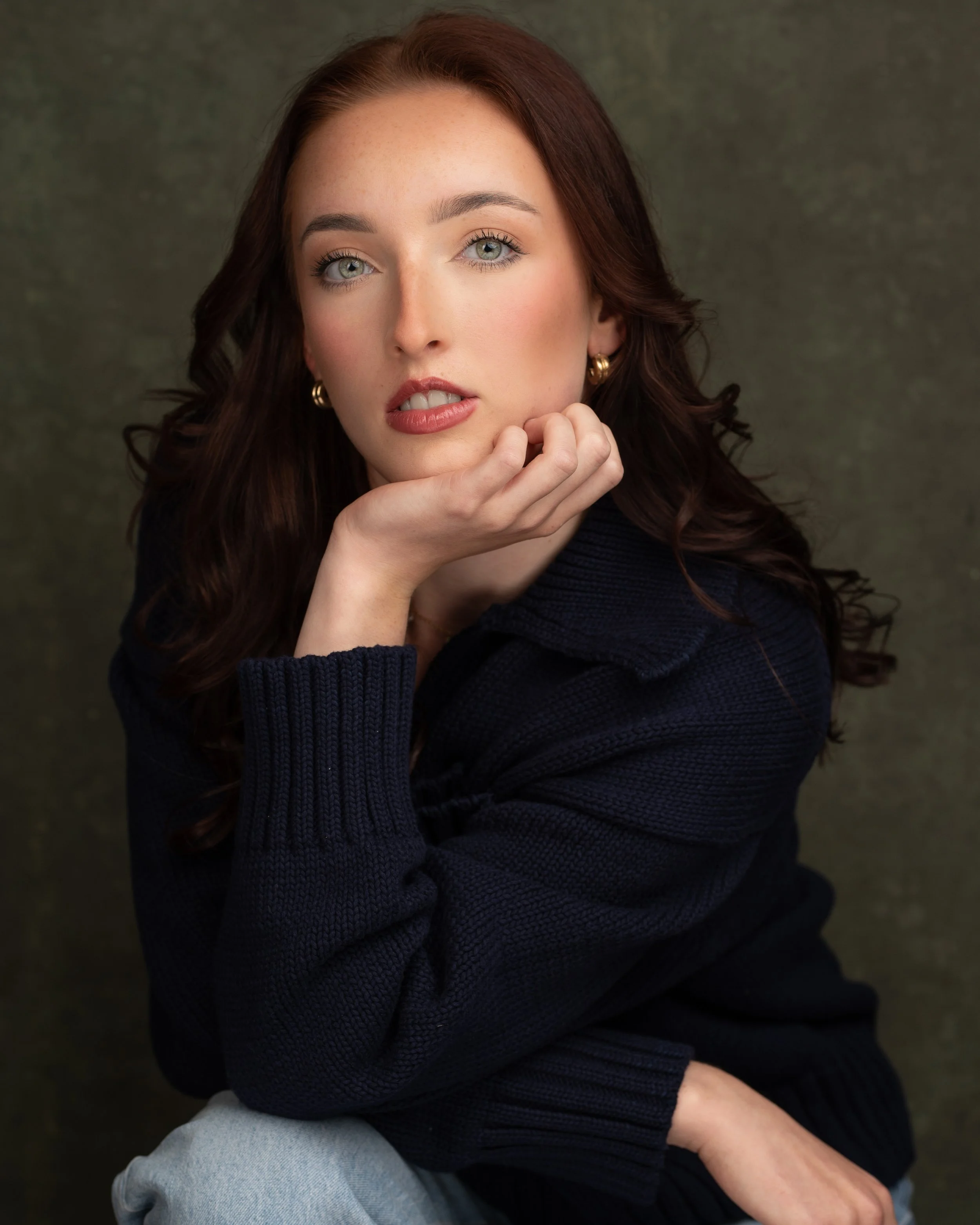 Portrait of a woman with red hair, blue eyes, wearing a dark sweater and gold hoop earrings, resting her chin on her hand, against a textured dark background.