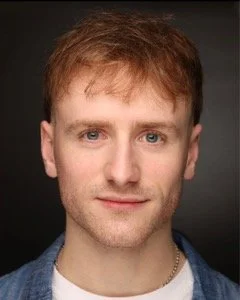 A young man with light skin, short reddish-brown hair, blue eyes, and a slight smile, wearing a denim jacket over a white t-shirt against a dark background.