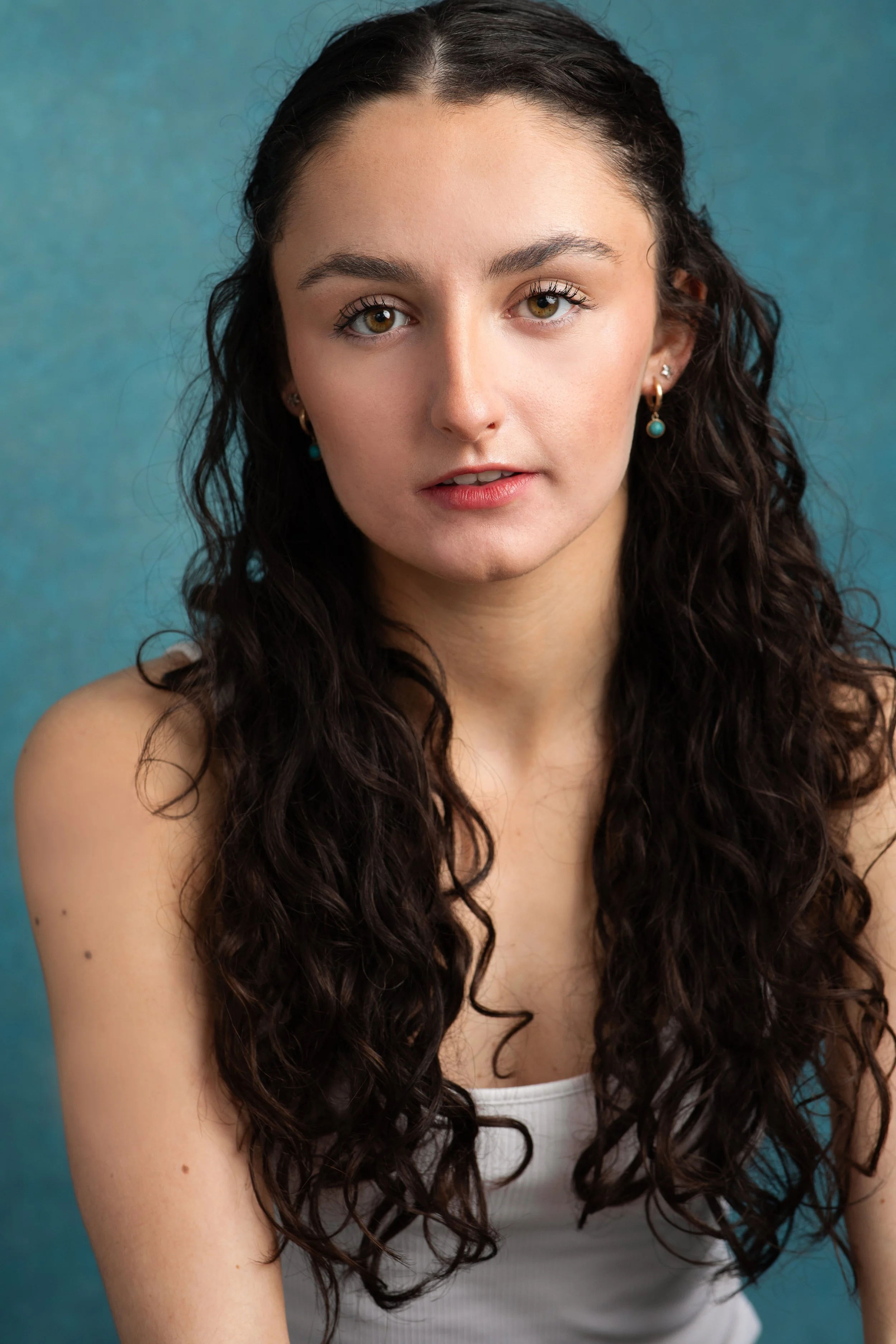 Portrait of a young woman with long, curly dark hair, wearing earrings, and a white sleeveless top against a blue background.