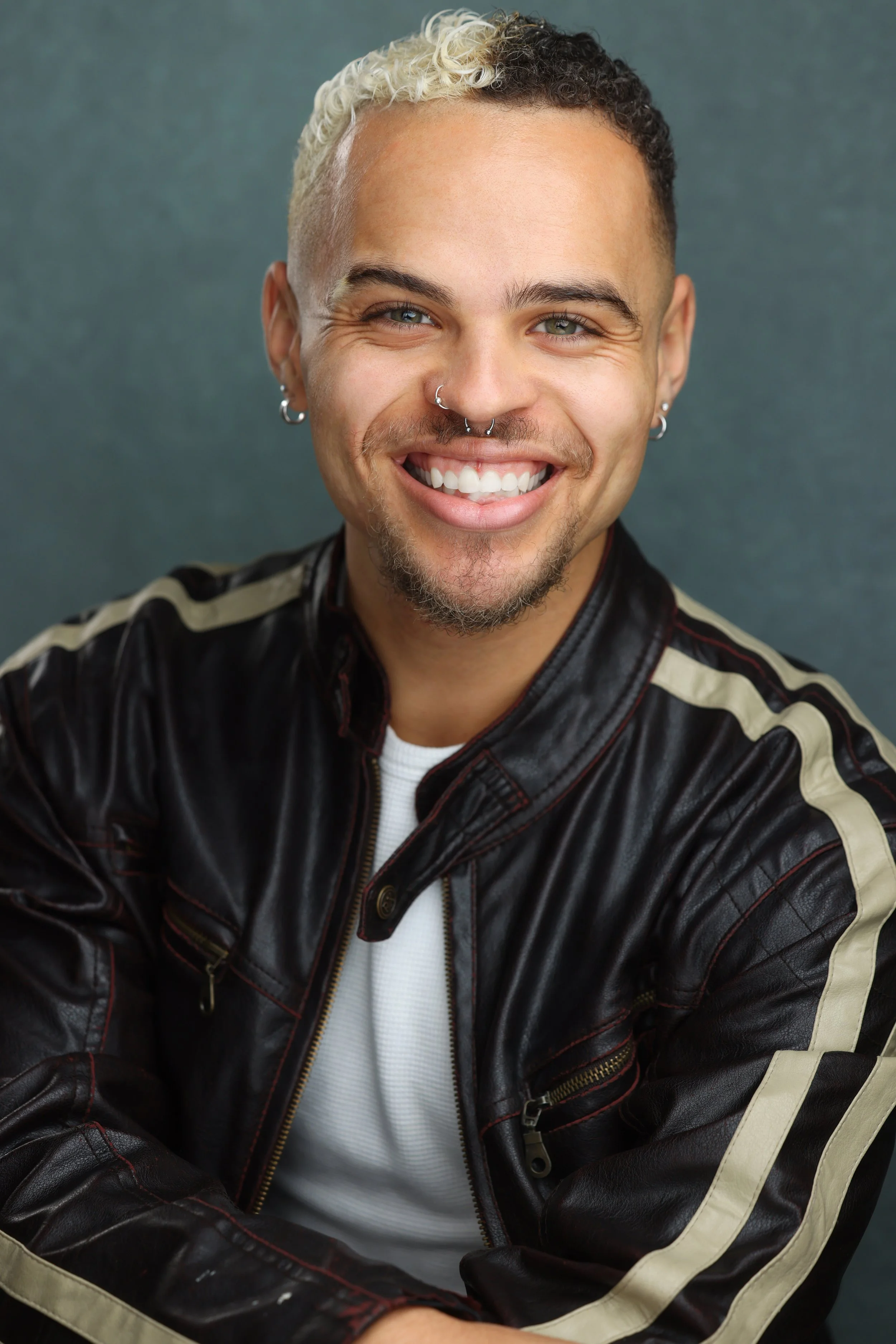 A young man with curly hair, multiple facial piercings, and a goatee, smiling in front of a dark background, wearing a black leather jacket with beige stripes and a white shirt.
