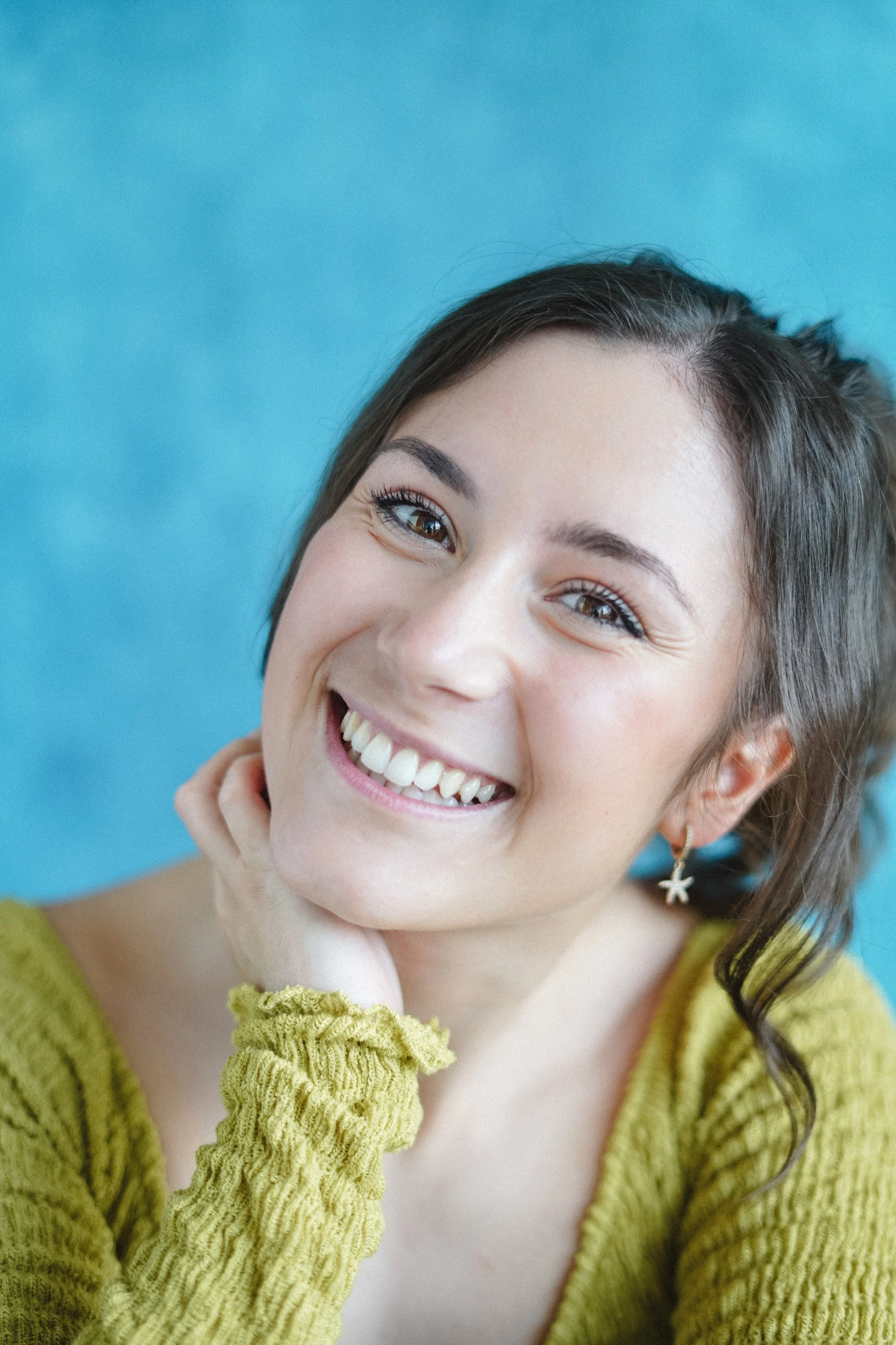 A young woman with brown hair and brown eyes smiling brightly, wearing a yellow-green sweater, with an earring shaped like a starfish, against a blue background.