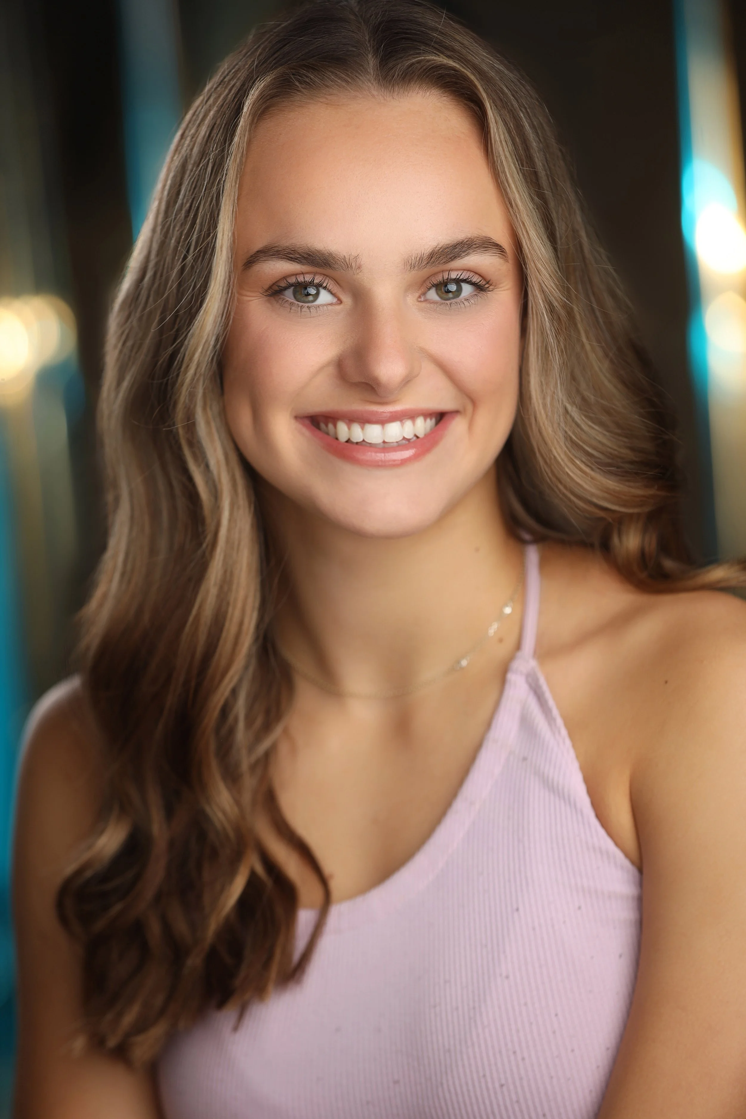 A young woman with light brown, wavy hair and hazel eyes smiling, wearing a purple tank top.