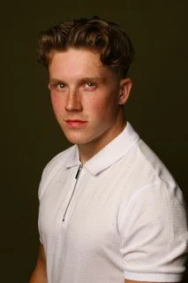 Portrait of a young man with light brown hair wearing a white collared shirt against a dark green background.