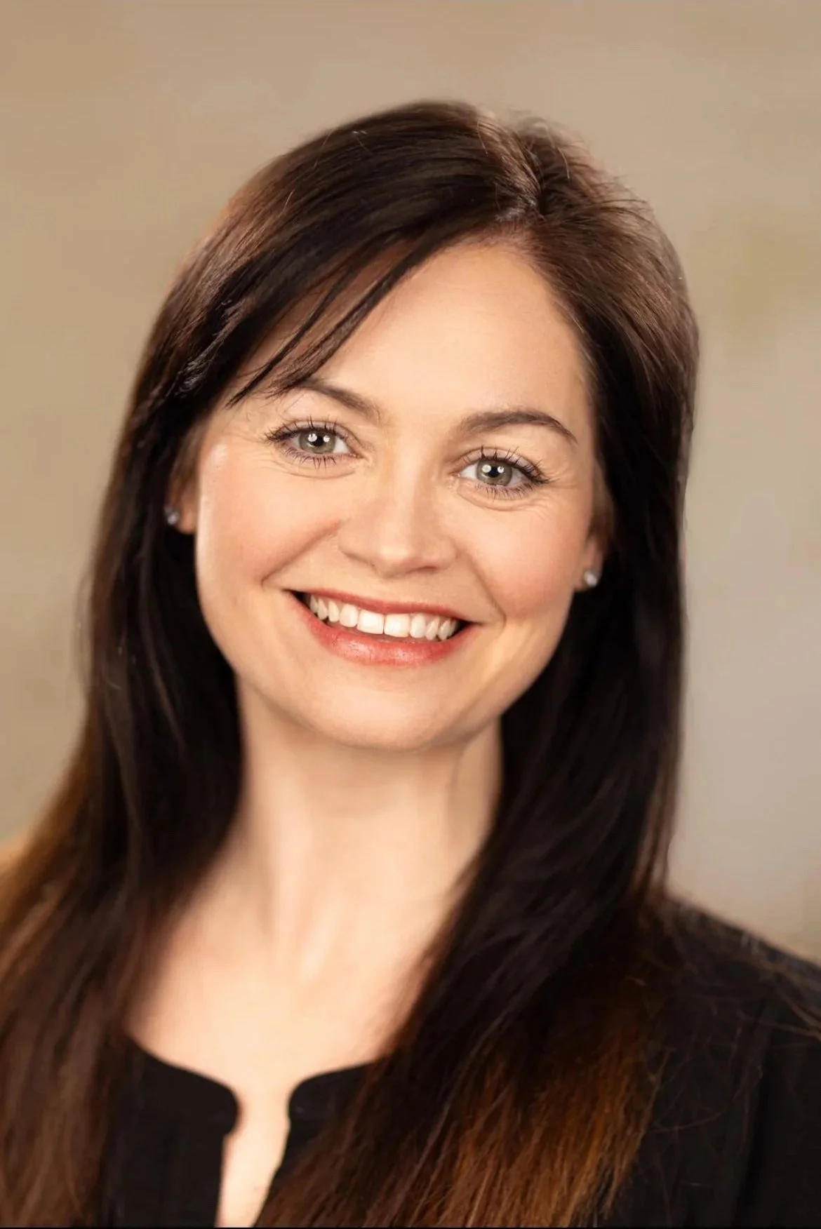 A woman with long brown hair and light eye color, smiling with white teeth, wearing a black top and small earrings, against a neutral background.