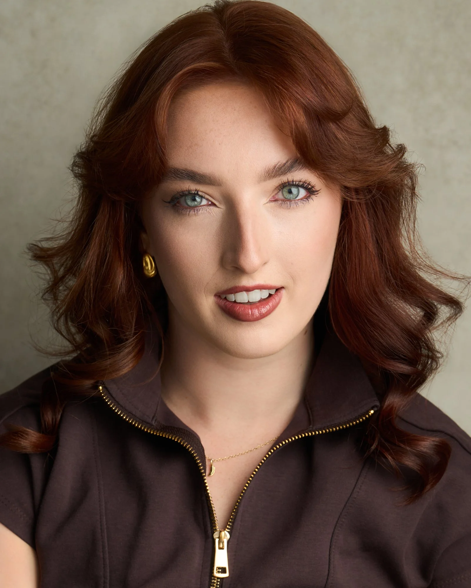 Portrait of a woman with red hair, blue eyes, wearing a dark sweater and gold hoop earrings, resting her chin on her hand, against a textured dark background.