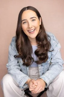 A woman with long brown hair and a bright smile, wearing a light denim jacket and sitting against a plain background.
