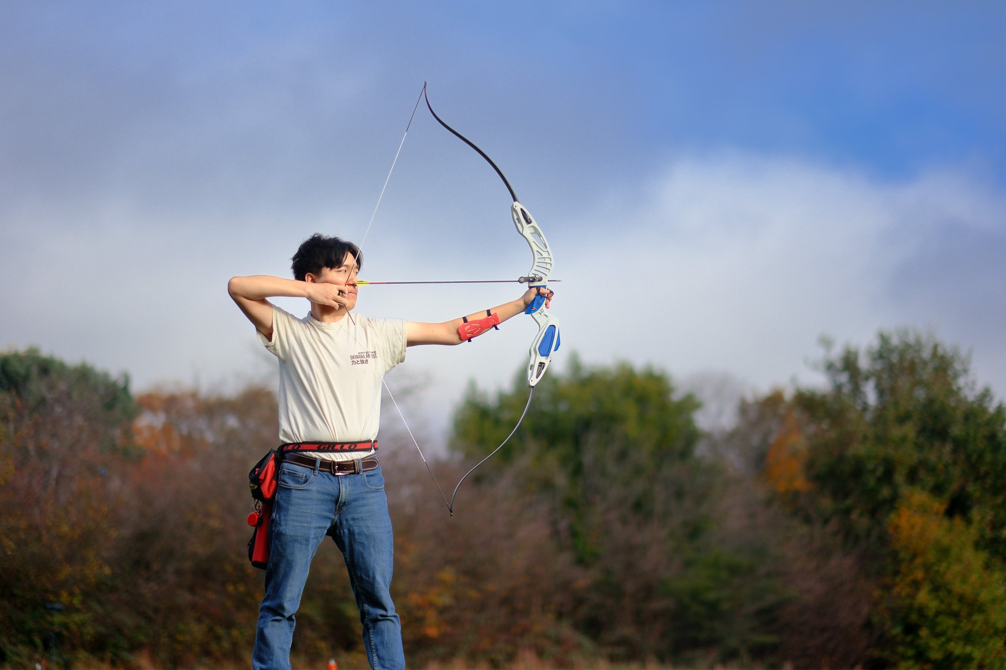 A young man outdoors practicing archery, drawing a bow with a arrow aimed at a target. He is wearing a light-colored t-shirt and blue jeans.
