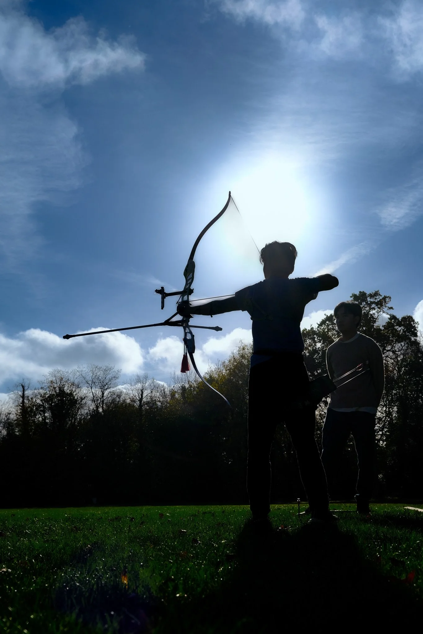 A person practicing archery outdoors during daytime, with another person, the archery coach watching, against a sky with clouds and the sun.