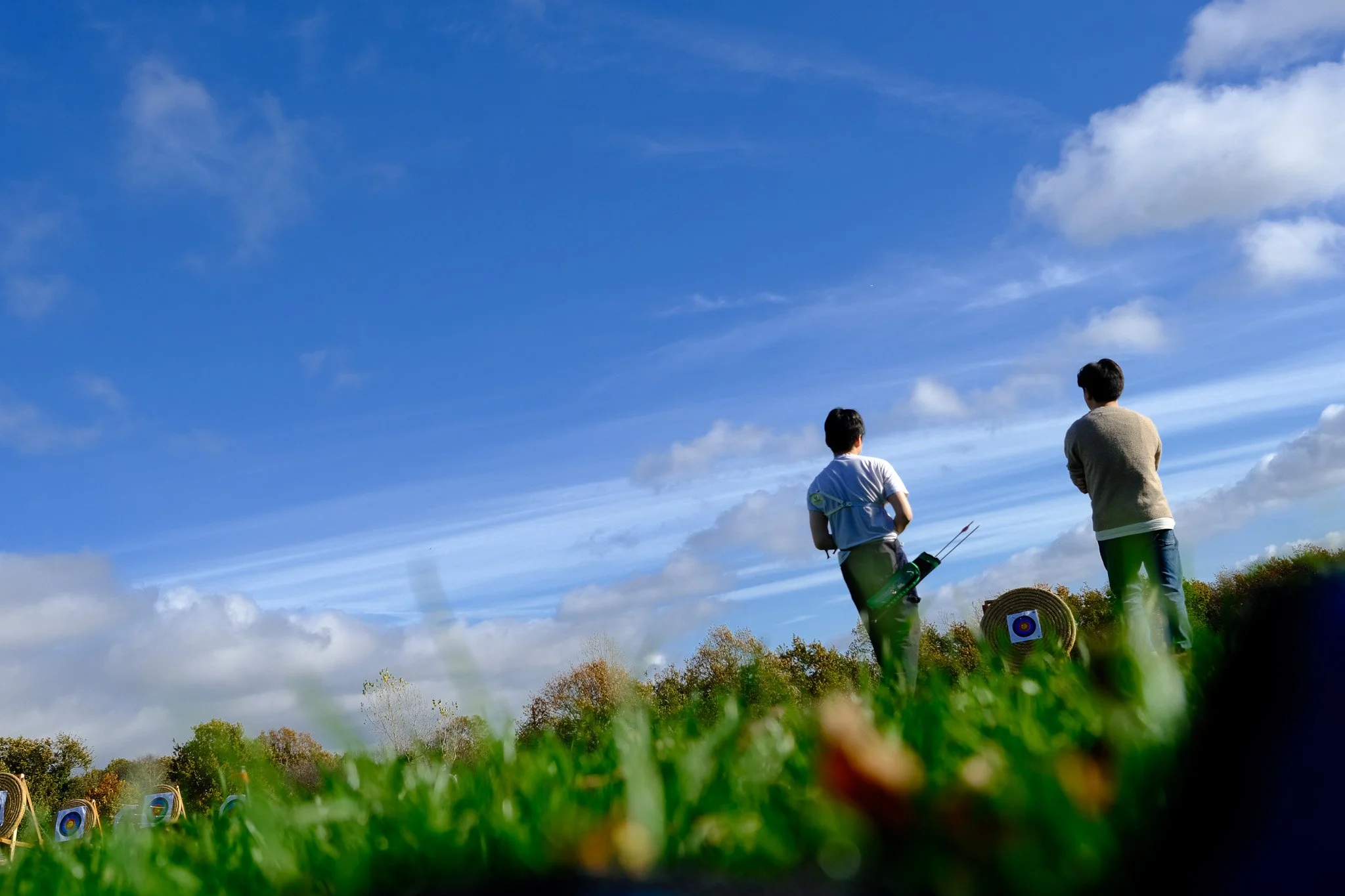 Two people standing on the grass at an outdoor archery range, facing away from the camera, walking towards their target, with a blue sky and some clouds overhead. There are targets set up in the distance.