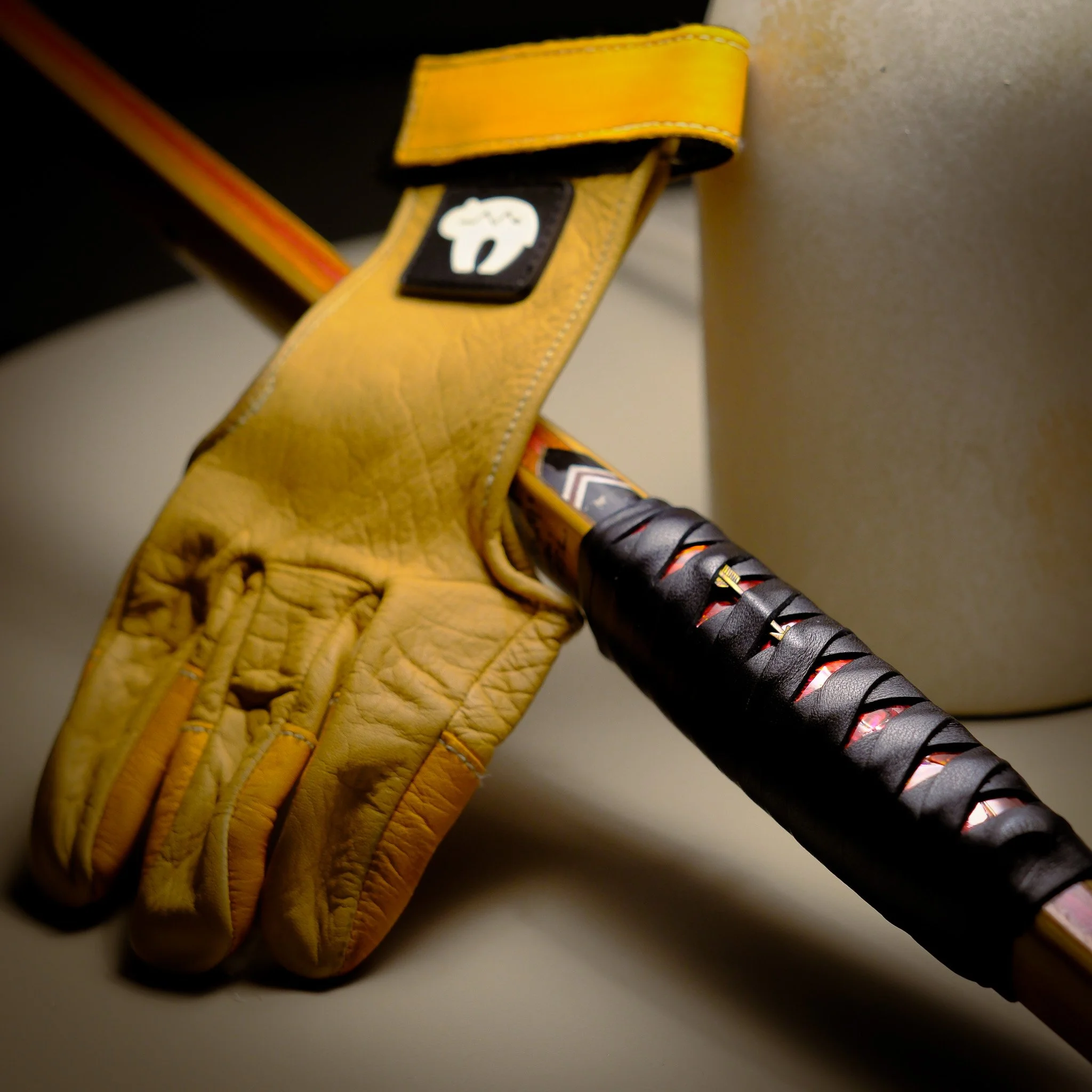 A yellow archery glove resting on a traditional English Longbow, wrapped in black tape beside it, simulating a katana look