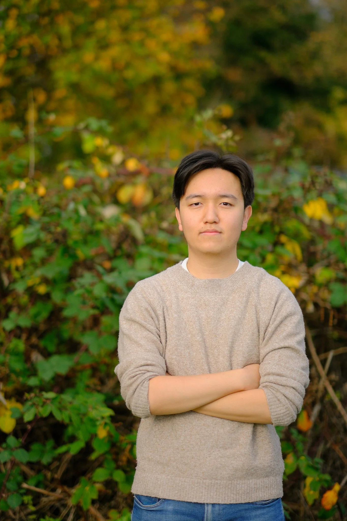A young man with black hair standing outdoors with arms crossed in front of a background of autumn foliage.