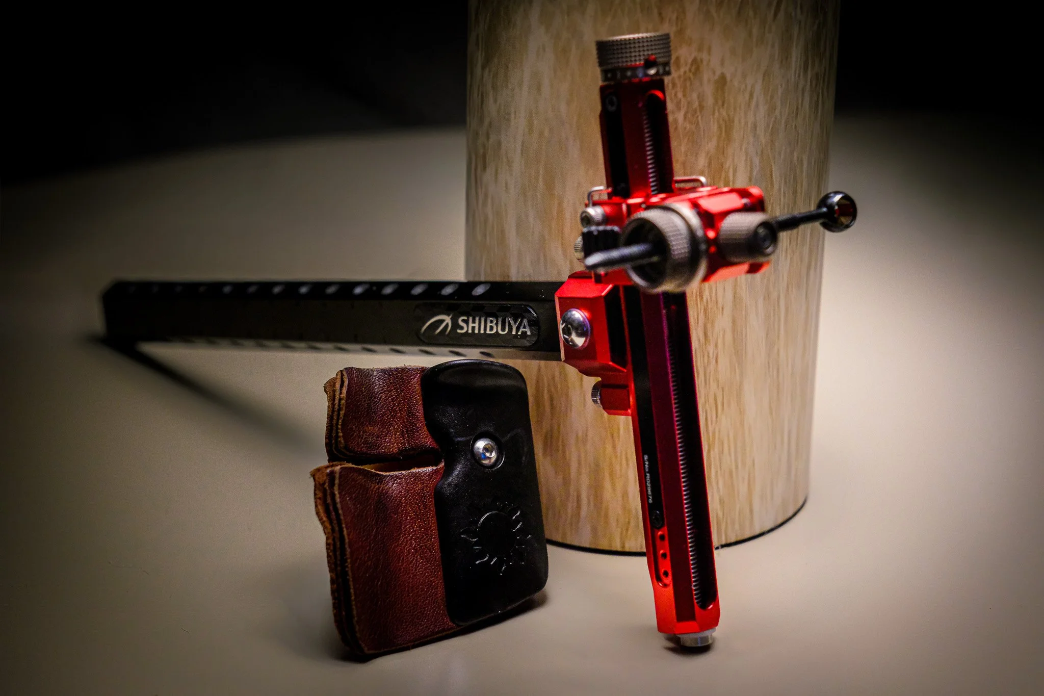 A red and black adjustable sight, leaning against a wooden cylinder, with a black and brown finger tab nearby.