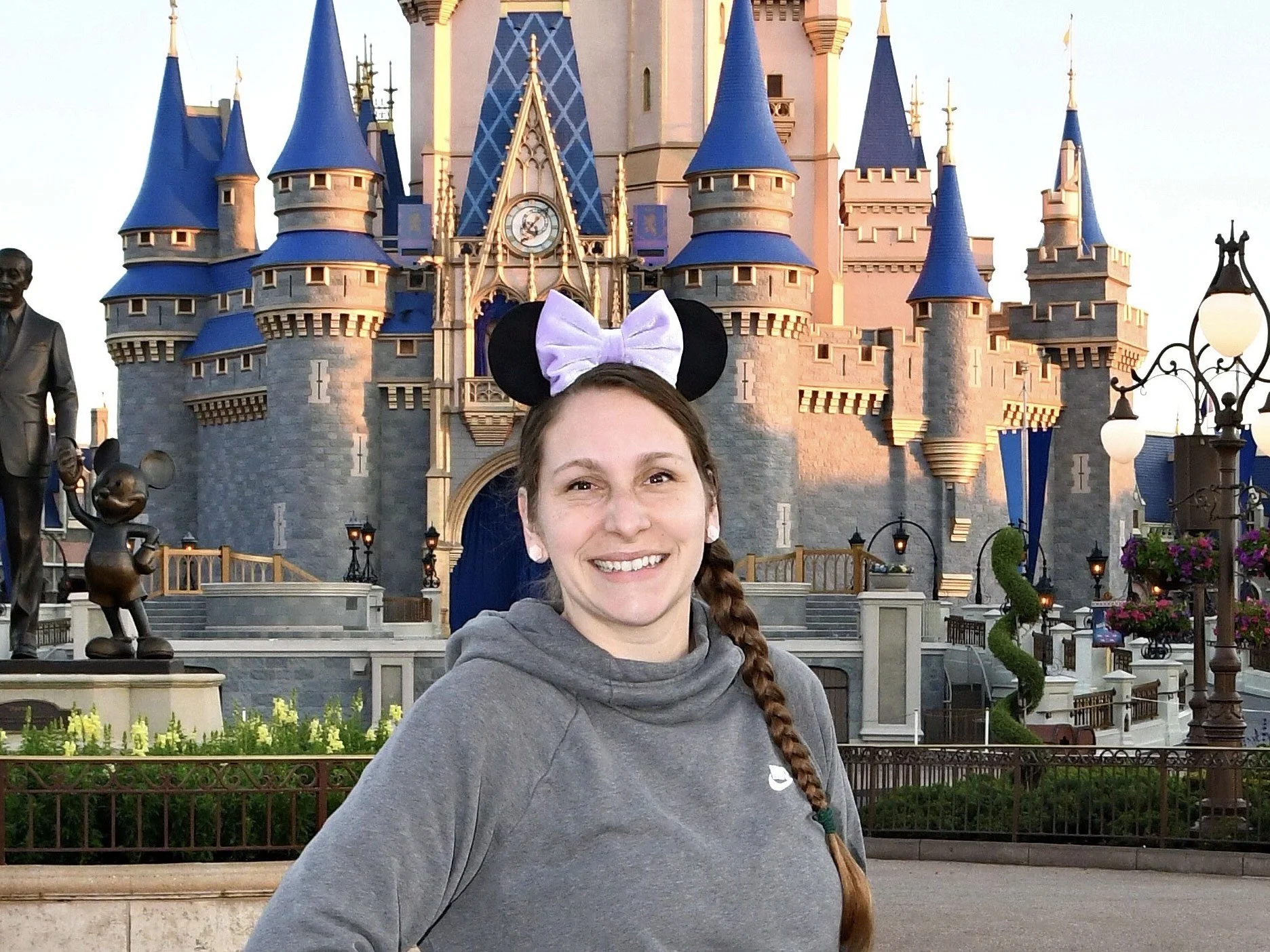 A woman with a braid, wearing a gray sweatshirt and Minnie Mouse ears, smiling in front of Sleeping Beauty Castle at Disneyland.