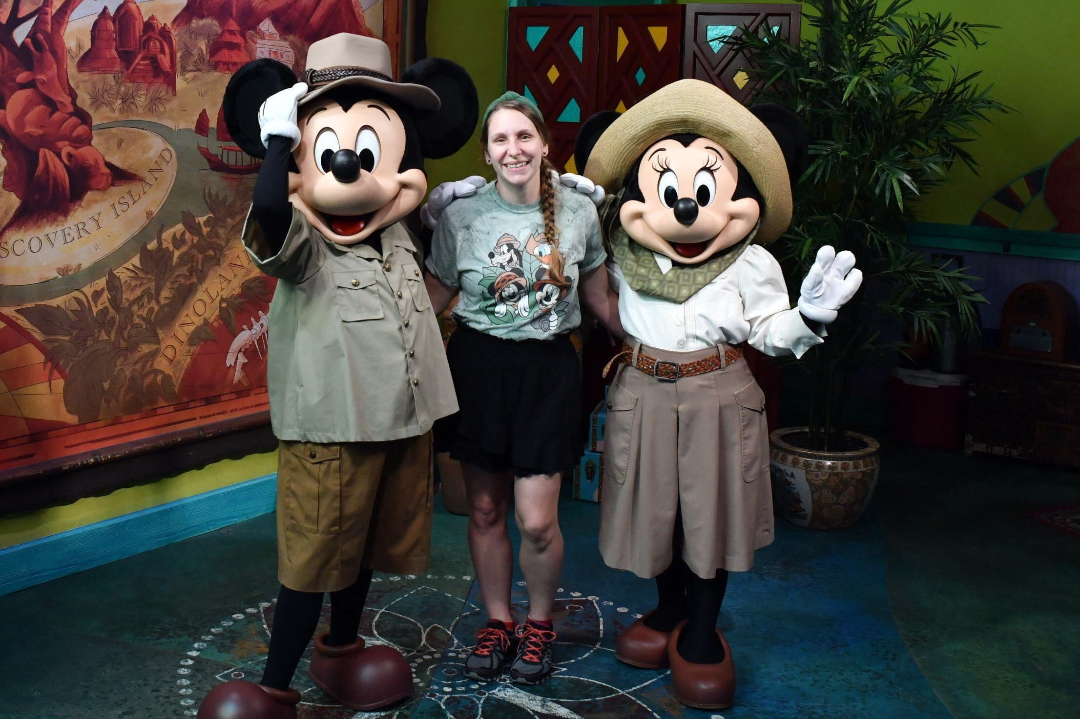 A woman standing between Mickey Mouse and Minnie Mouse, all three smiling and waving in a colorful indoor setting with jungle-themed decorations.