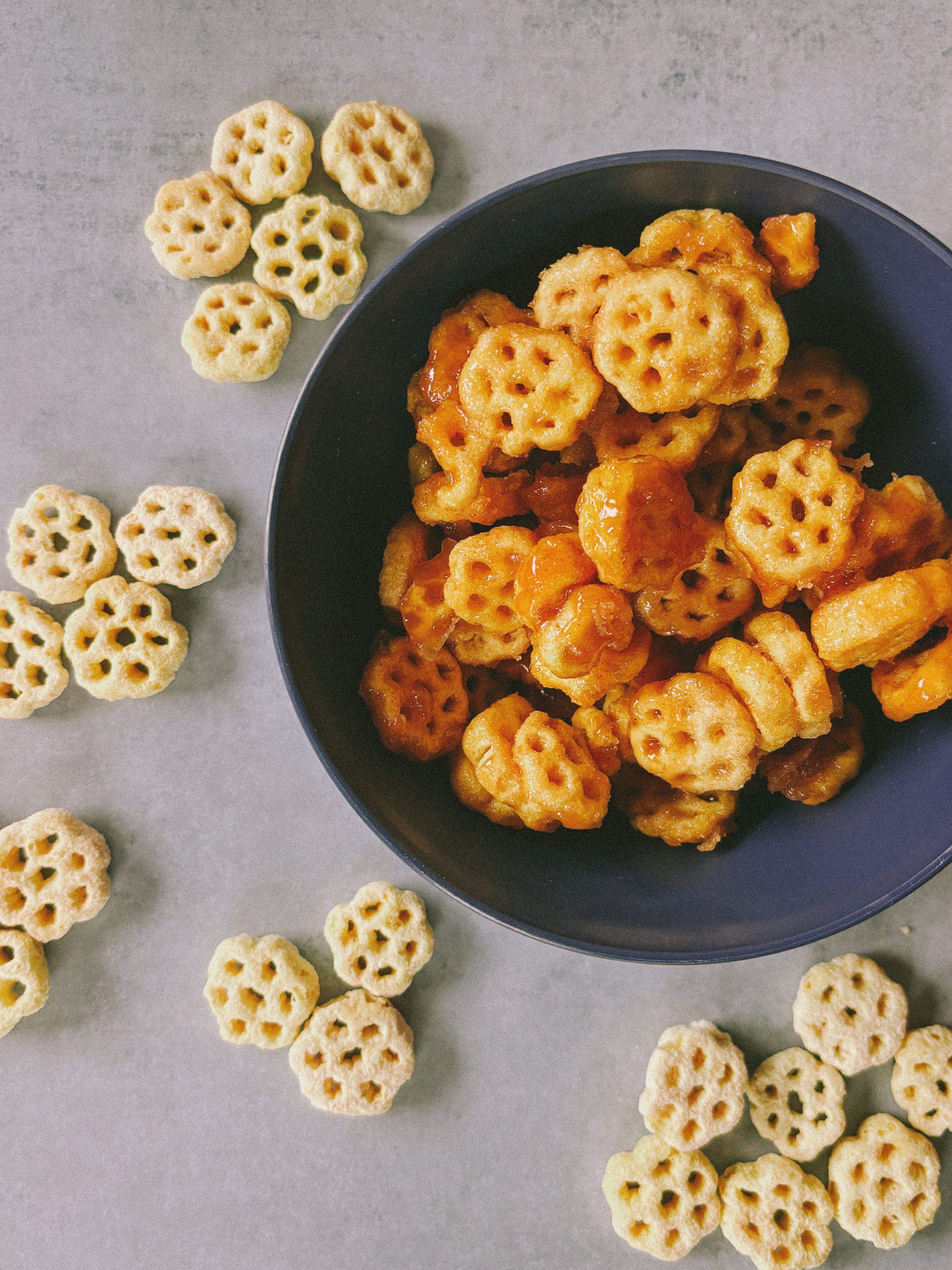 A black bowl filled with honey-glazed mini waffle cookies, surrounded by scattered similar cookies on a light gray surface.