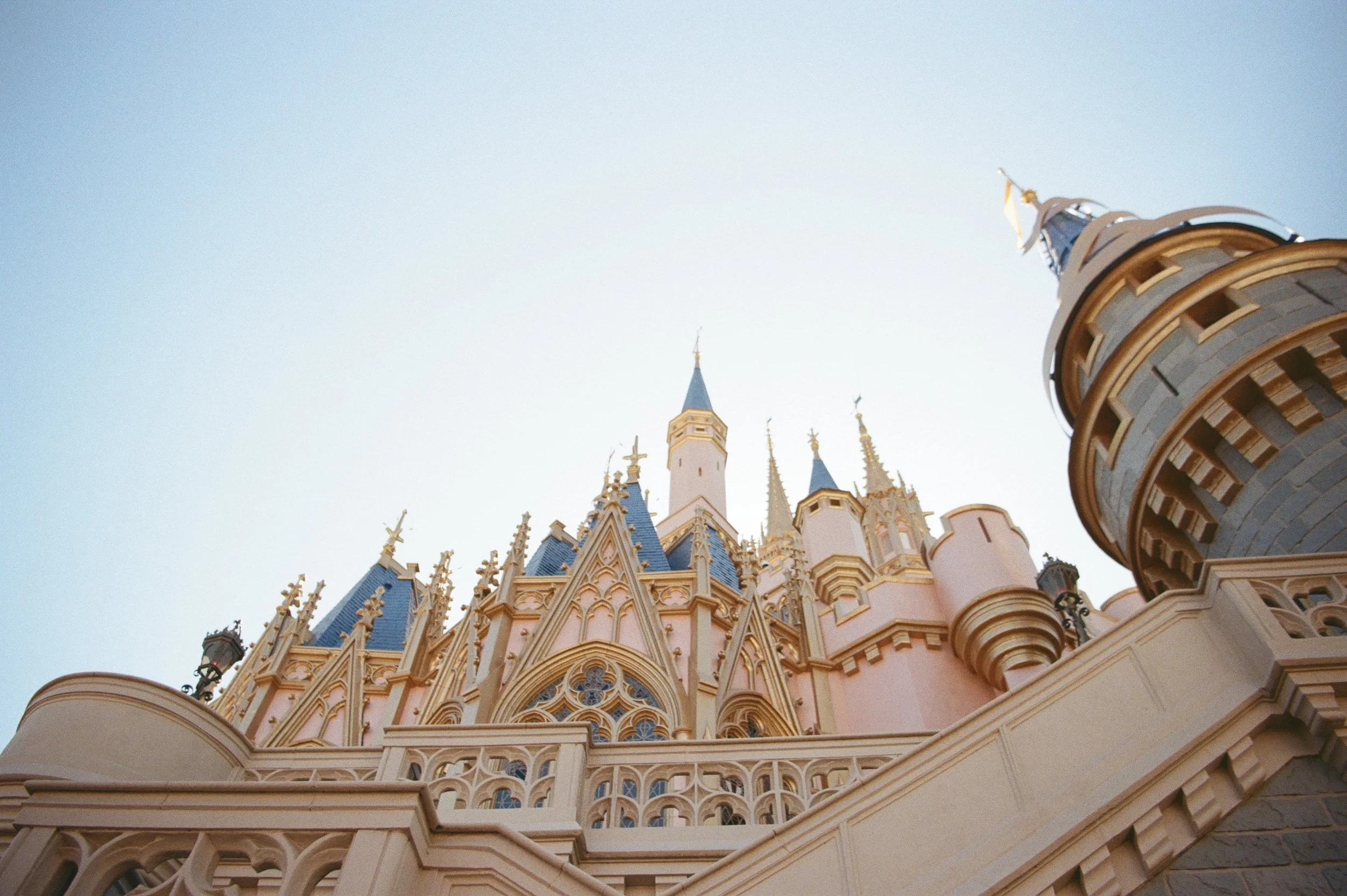 Looking up at a fairy tale castle with pink walls, blue roofs, and golden accents against a pale sky.