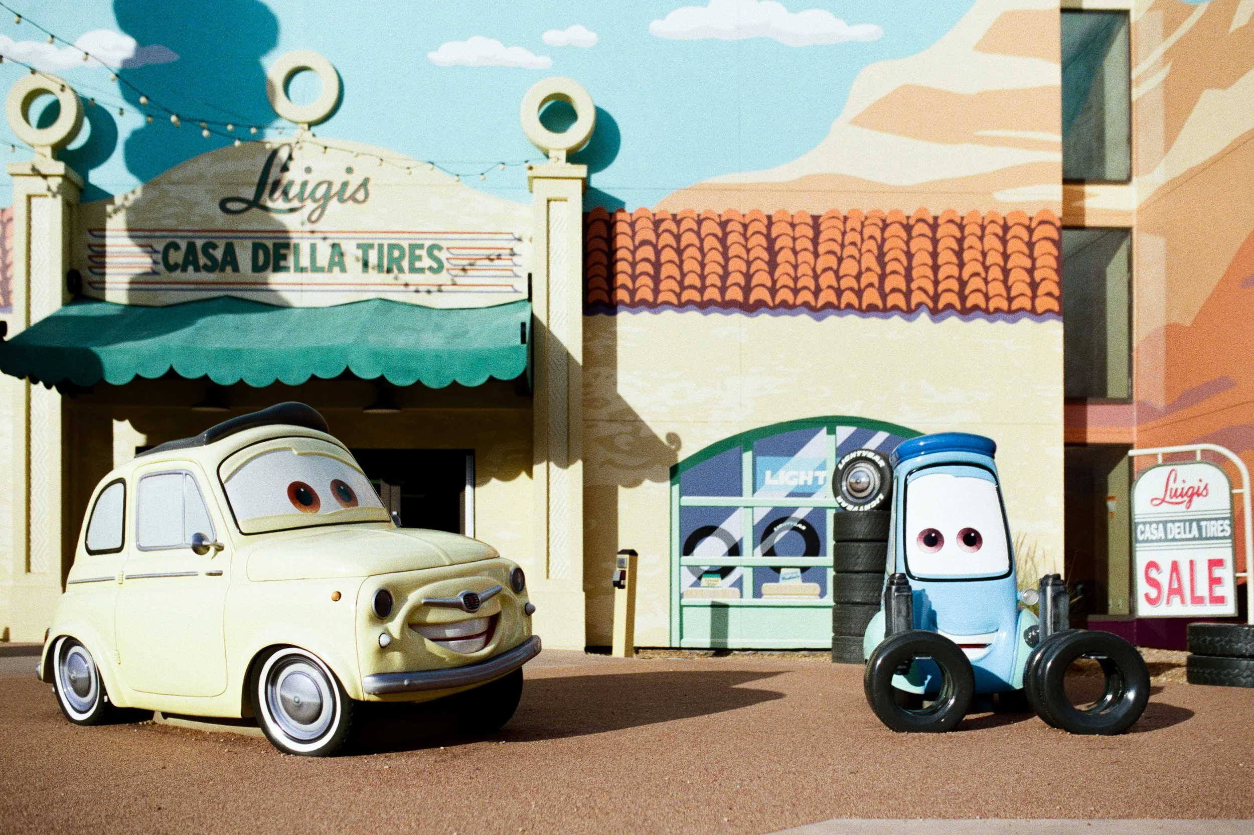 Animated scene featuring two cartoon cars with eyes and smiles outside an auto repair shop named 'Luigi's Casa Della Tires,' with a garage sale sign and shelves of tires in the background.