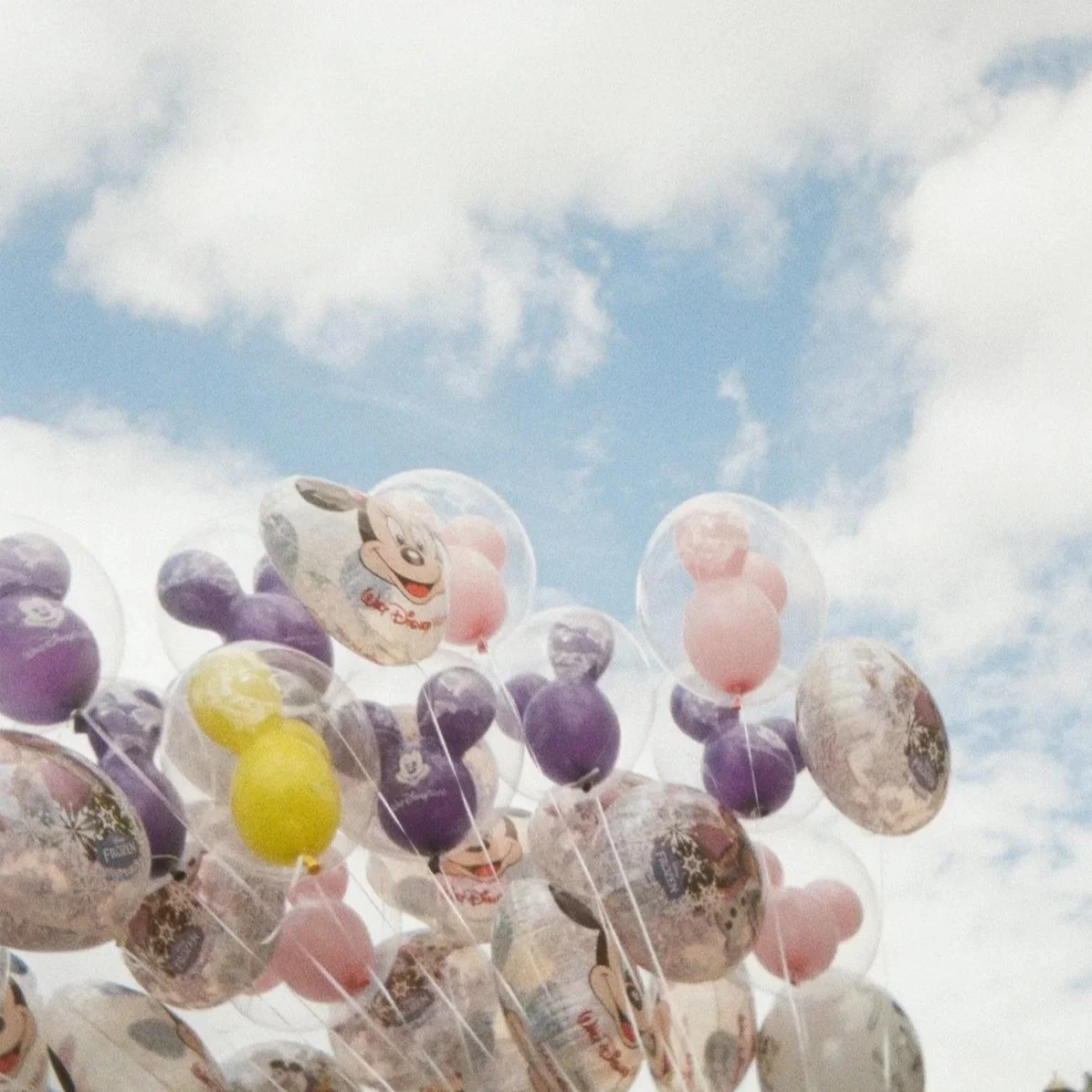 A cluster of colorful balloons, including ones with Mickey Mouse images, floating against a cloudy sky.