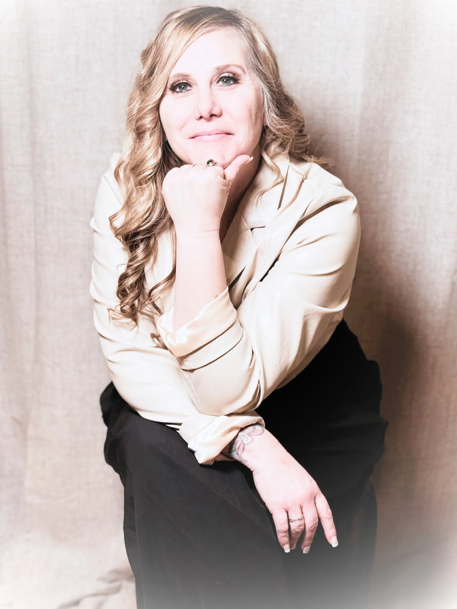 A woman with long, wavy blonde hair, wearing a cream-colored satin blouse and black pants, sitting with her chin resting on her hand, smiling softly at the camera against a beige textured background.