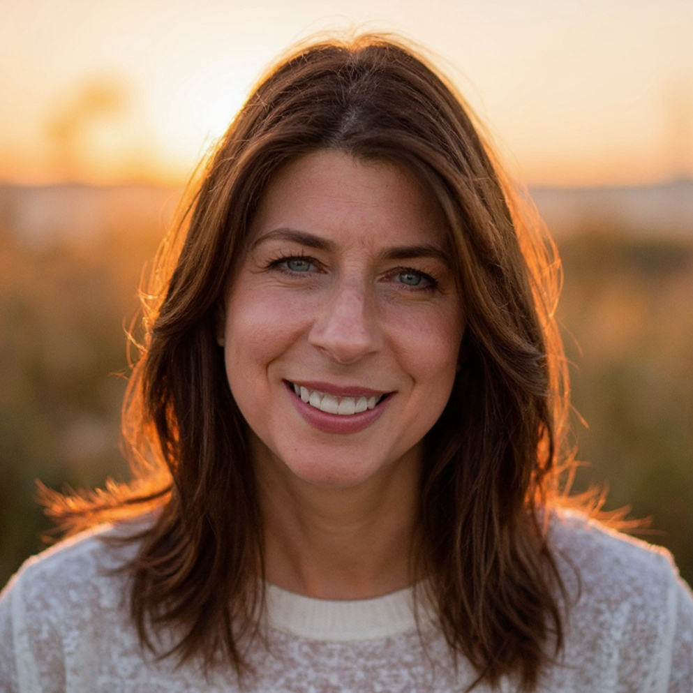 Close-up portrait of a smiling woman with brown hair and blue eyes, outdoors at sunset with blurred background.