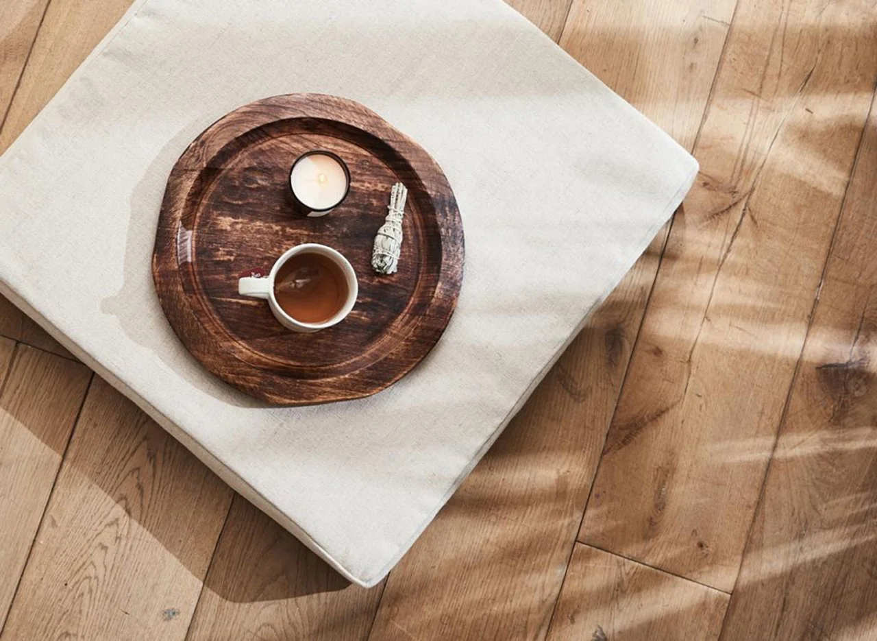 A wooden tray with a cup of tea, a lit candle in a holder, and a bundle of sage, placed on a white cloth on a wooden floor.