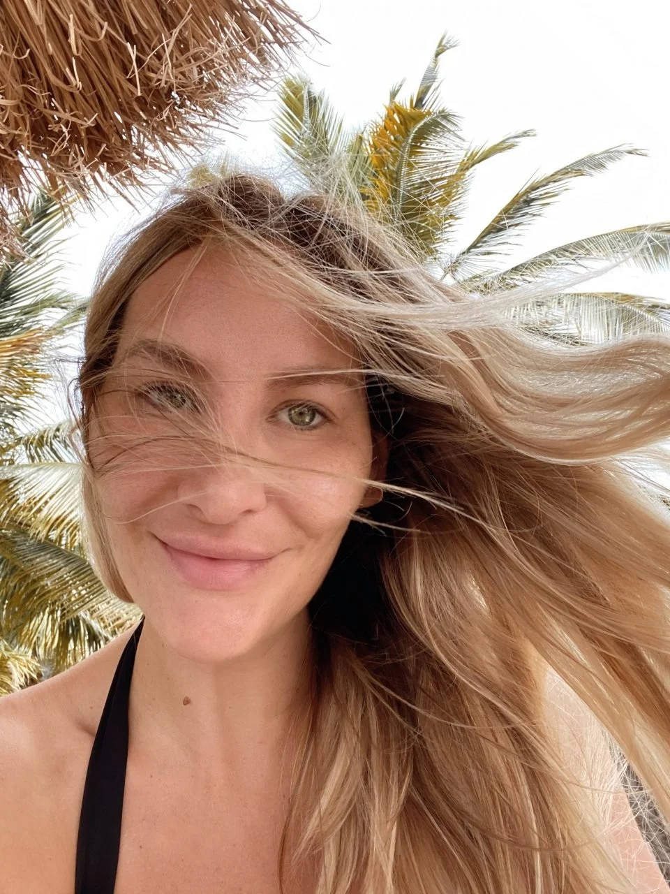 A woman with long, blonde hair partially covering her face, smiling, with palm trees and a thatched umbrella in the background on a sunny day.