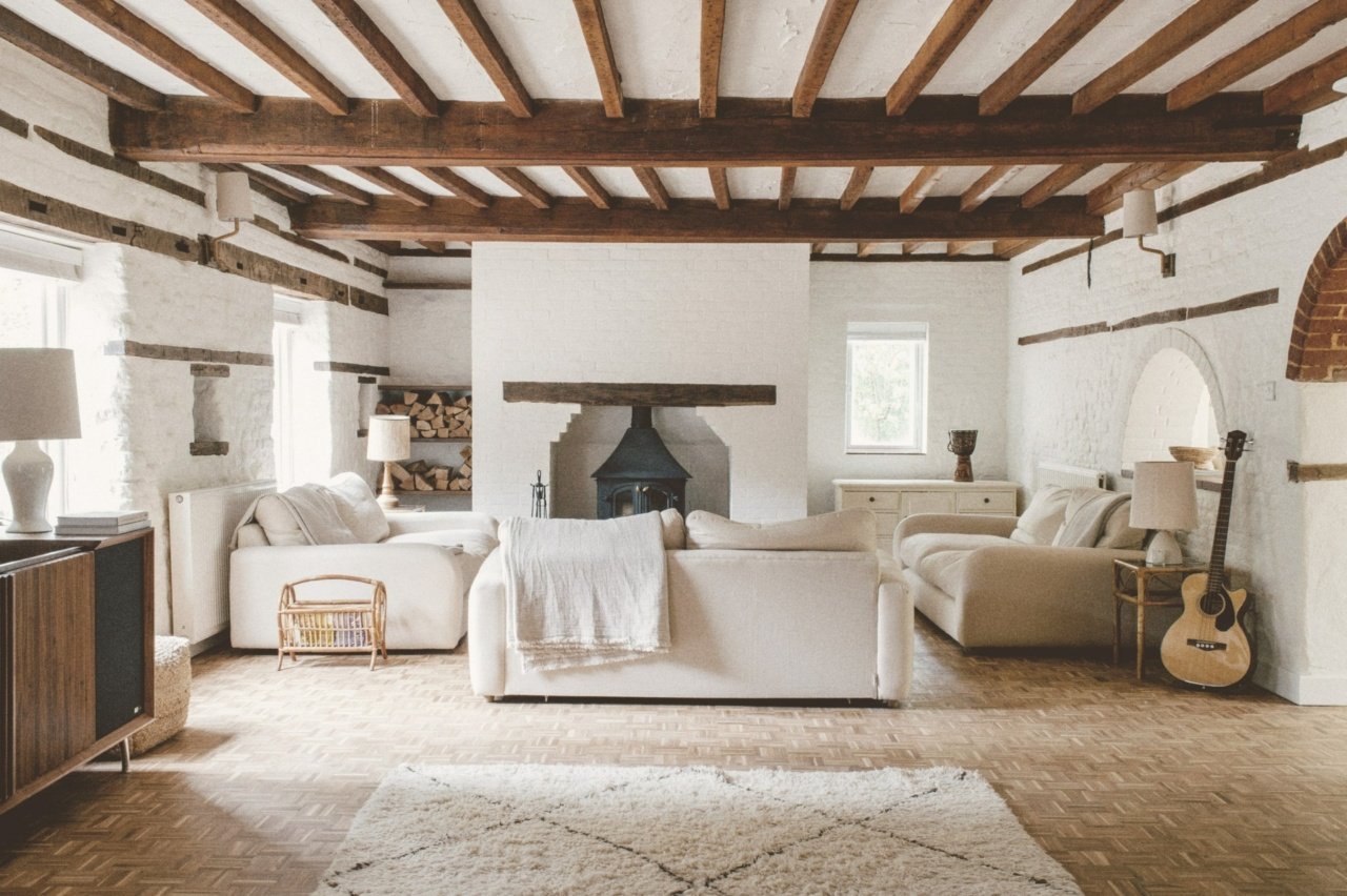 Living room with white sofas, wooden ceiling beams, a black fireplace, a guitar, side tables with lamps, and a white rug.