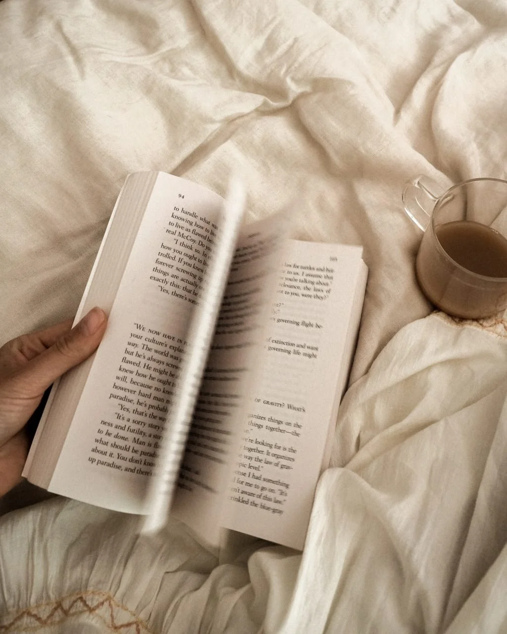 A person holds an open book on a white bed with crumpled sheets, next to a glass of coffee or tea.
