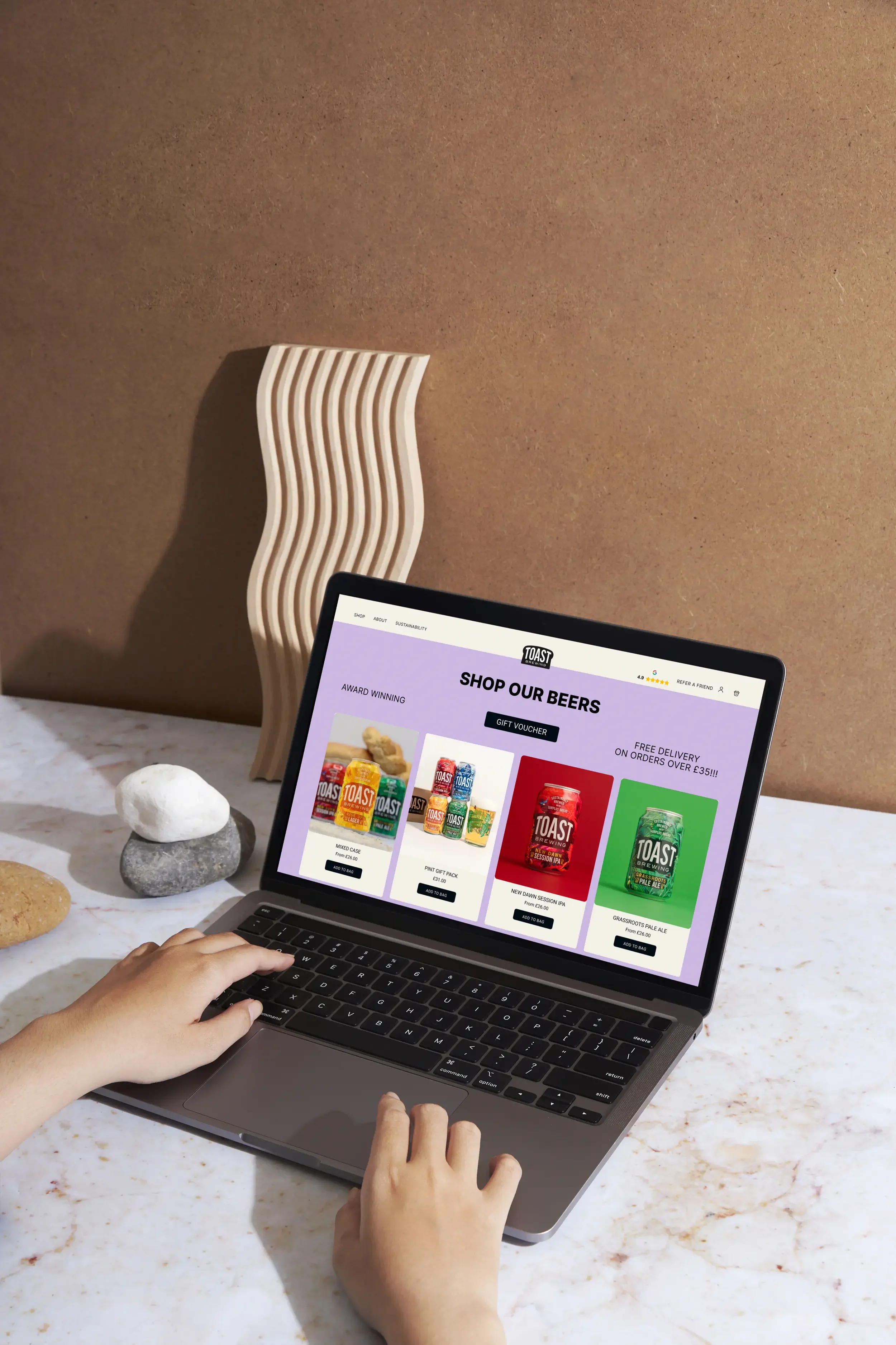 Person browsing an online shop for beers on a laptop, with rocks and a sculptural wooden object on a marble surface.