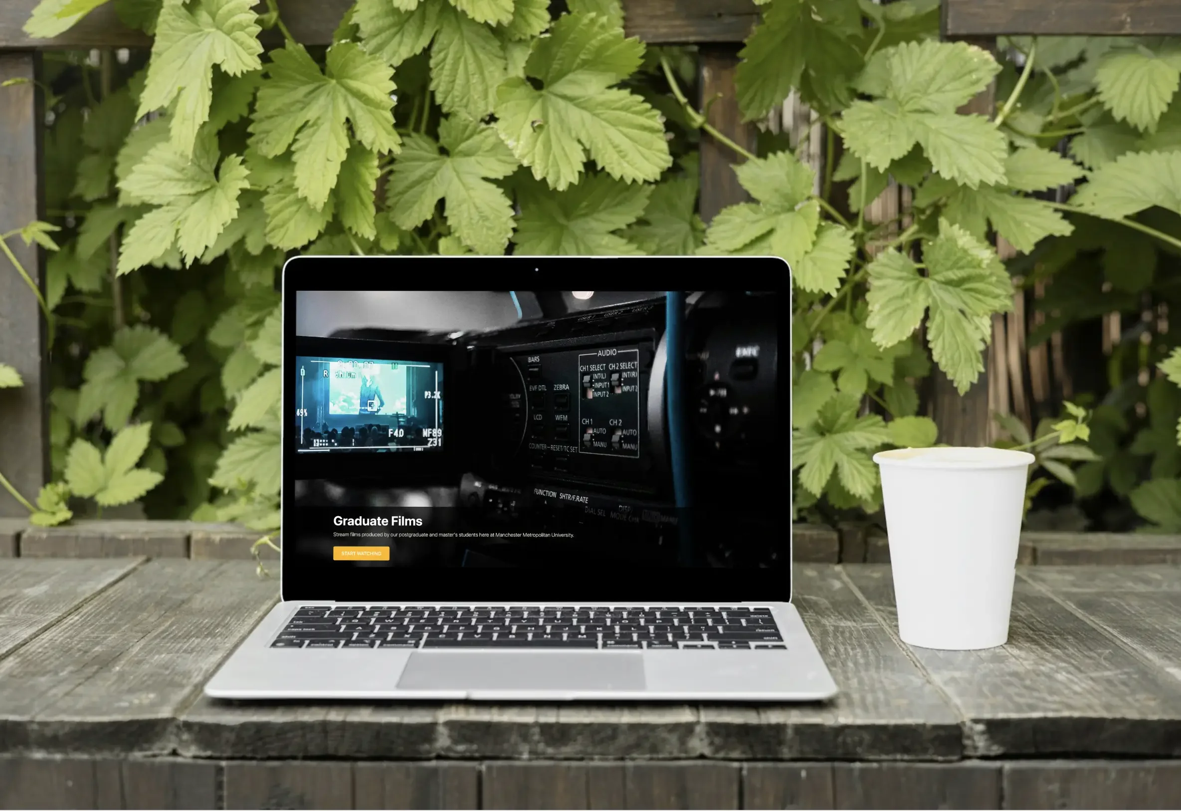 Laptop displaying a video on a wooden table outdoors, with a white paper cup to the right and green leafy plants in the background.